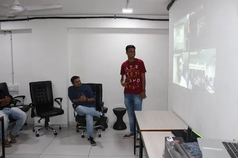 A man stands beside a seated man in an office room while a projector screen shows a grid of video thumbnails.