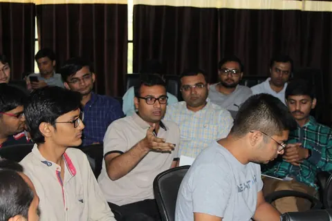 A man in a beige polo shirt gestures while speaking among rows of seated attendees at a meetup.
