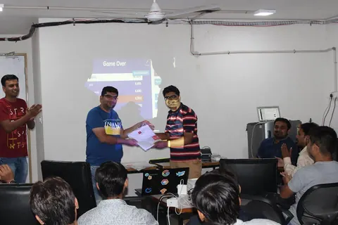 Two men exchange a folded white t-shirt in front of a projected Game Over leaderboard as attendees clap.