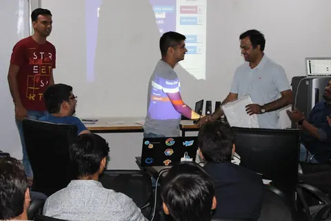 A man shakes hands with another holding a white envelope as a results screen is projected behind them.