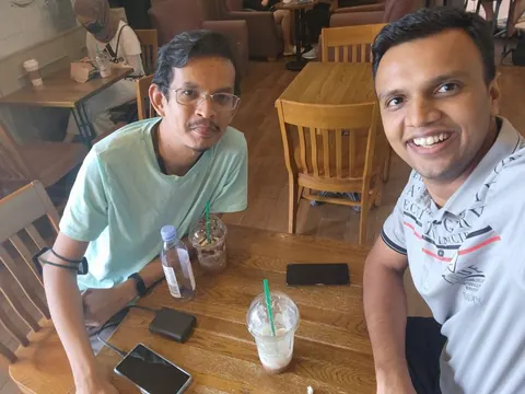 Two men smile in a cafe selfie beside iced drinks, a water bottle, smartphones and a power bank.