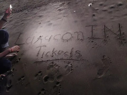 Person crouching on a beach at night writes 'Laracon IN Tickets' in the sand, lit by a phone flashlight.