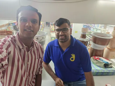 Two men smile in a selfie inside a supermarket aisle with stocked shelves and stacked housewares behind them.