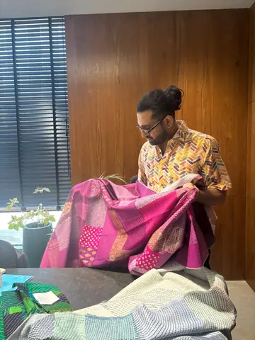 A man in a patterned shirt examines a magenta patchwork textile at a table stacked with folded green and white fabrics.