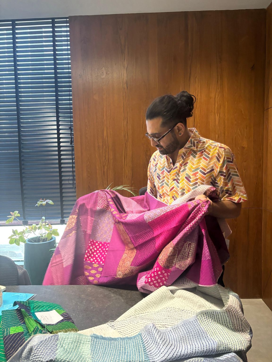 A man in a patterned shirt examines a magenta patchwork textile at a table stacked with folded green and white fabrics.