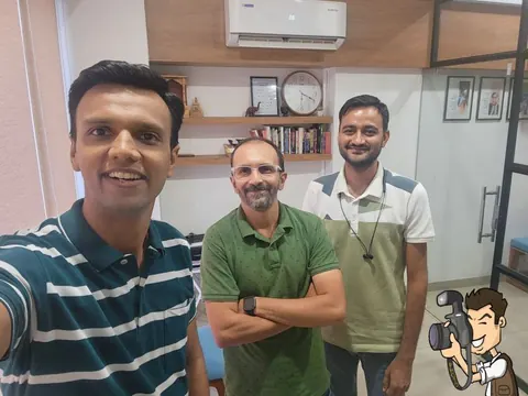 Three men smile for a selfie in an office with a wall clock, bookshelf and air conditioner behind them.