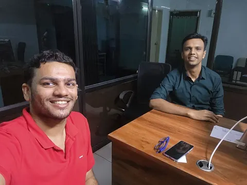 Two men smile in a selfie taken in an office, one seated at a wooden desk with a phone and glasses on it.