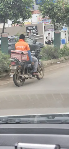 Delivery rider in an orange Swiggy t-shirt on a motorcycle carrying a pink Zomato delivery bag on a city street.