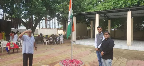 A man salutes the Indian tricolour as two men stand nearby during an outdoor flag ceremony.