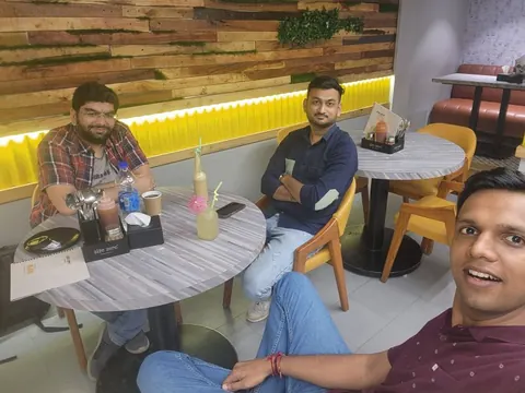 Three men at a coffee shop, the man in front taking a selfie, with drinks and menus on marble-top tables.