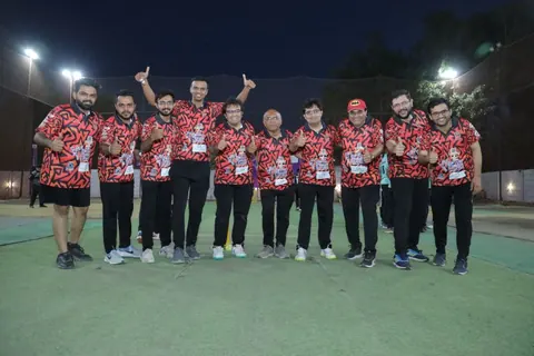 A group of men in matching red and black jerseys pose with thumbs-up on a floodlit cricket pitch at night.