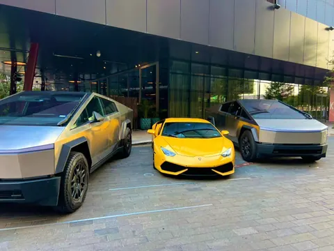 Two Tesla Cybertrucks parked either side of a yellow Lamborghini outside a modern glass building.