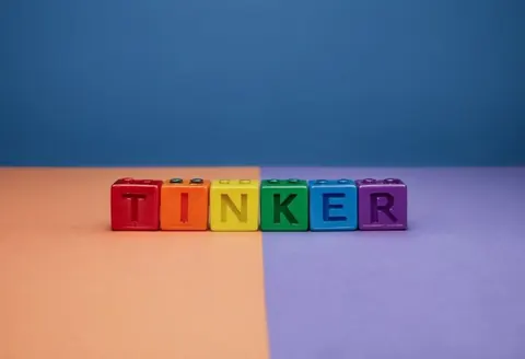Colourful letter blocks spelling TINKER on a peach and purple surface against a blue background.