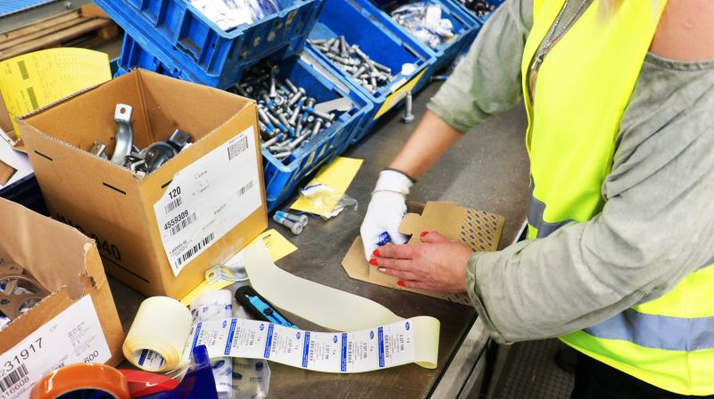 A woman in a high-visibility vest labels a small box at a workbench with bins of parts, tape and printed labels.