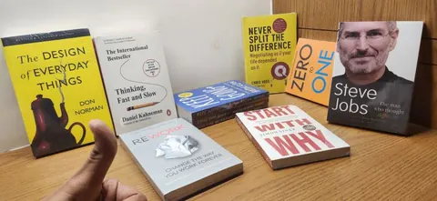 Thumb pointing at a display of books including Design of Everyday Things, Steve Jobs and Start with Why on a desk.