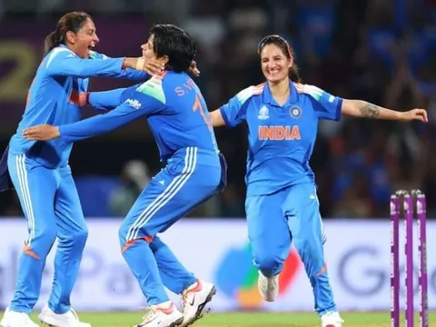 Three Indian women cricketers in blue jerseys celebrate on the field, one leaping mid-air with joy during a match.