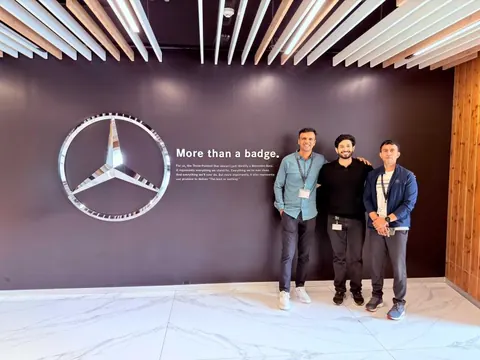 Three men pose beside a Mercedes-Benz star logo and 'More than a badge' wall display.