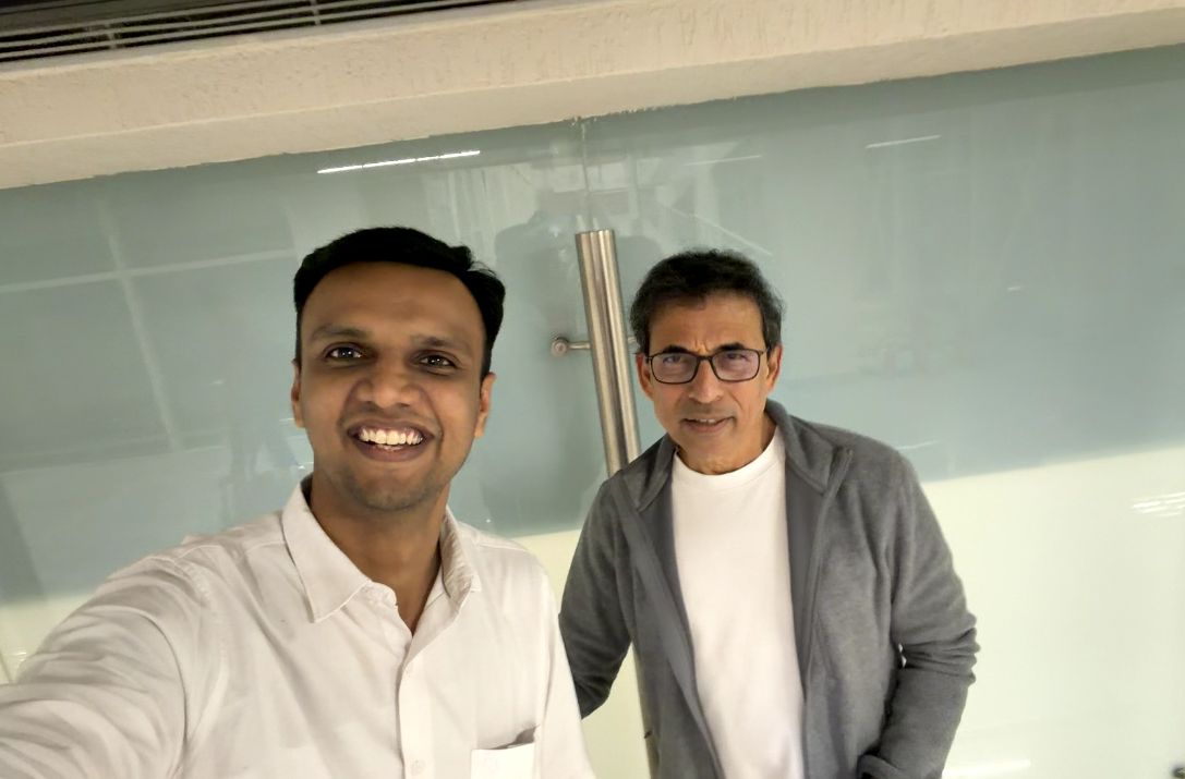 Gaurav smiles in a selfie beside Harsha Bhogle, who wears glasses, a grey jacket and white t-shirt.