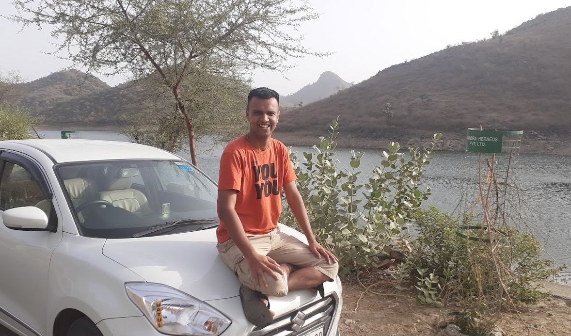 Gaurav sits cross-legged on the bonnet of a white car beside a lake, smiling, with dry hills and trees behind.