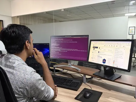 A man at an office desk reviews code on a terminal screen beside a monitor showing a social media feed.