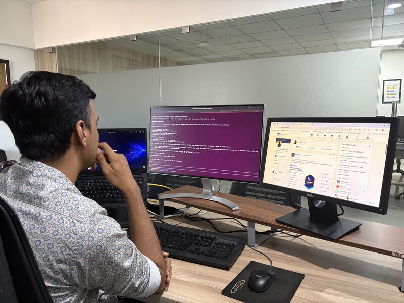 A man at an office desk reviews code on a terminal screen beside a monitor showing a social media feed.