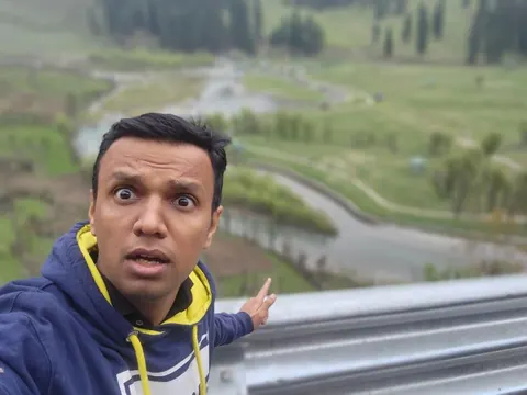 Gaurav pulls a surprised face in a selfie taken behind a guardrail above a green river valley.