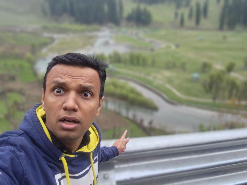 Gaurav pulls a surprised face in a selfie taken behind a guardrail above a green river valley.