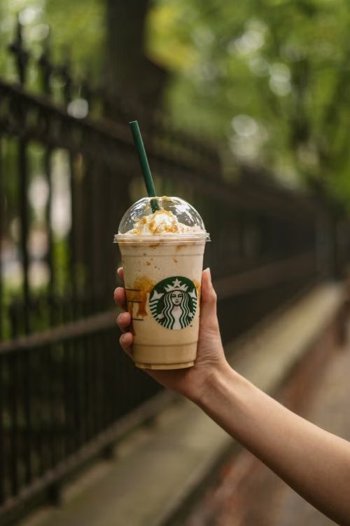 A hand holds a Starbucks iced coffee with whipped cream and a green straw in front of an iron fence.