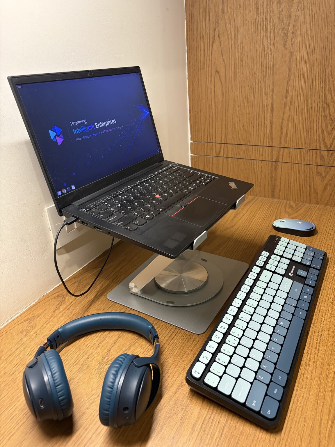 Laptop raised on a metal stand at eye level on a wooden desk, with a wireless keyboard, mouse and blue headphones below.