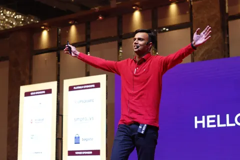 A speaker in a red polo shirt stands on stage, arms outstretched, before sponsor banners and a purple screen.
