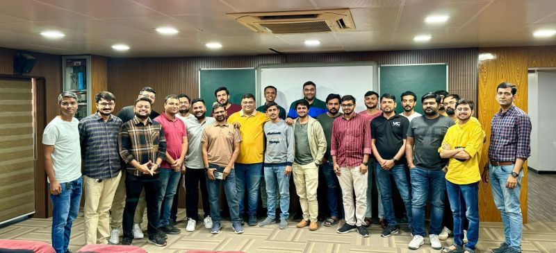 About twenty men pose for a group photo in a meeting room with green boards.