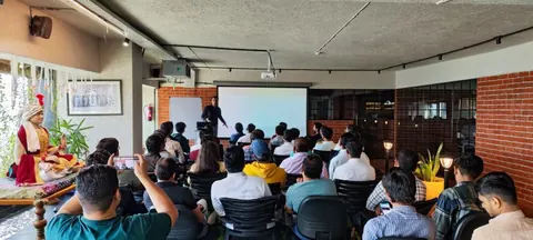 A speaker presents beside a projection screen to a seated audience in a brick-walled room.