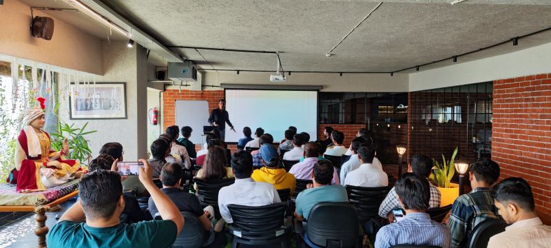 A speaker presents beside a projection screen to a seated audience in a brick-walled room.