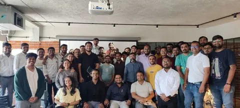 A large group of meetup attendees pose in a brick-walled venue with a projector.