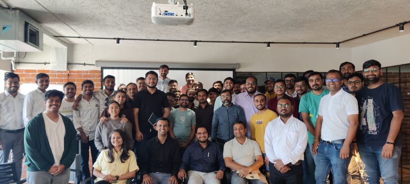 A large group of meetup attendees pose in a brick-walled venue with a projector.
