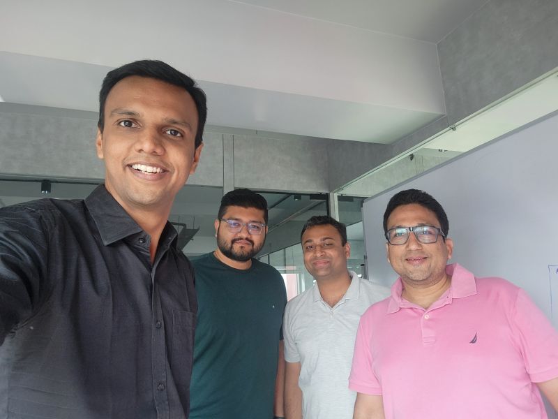 Four men smile in a selfie inside an office with a whiteboard.