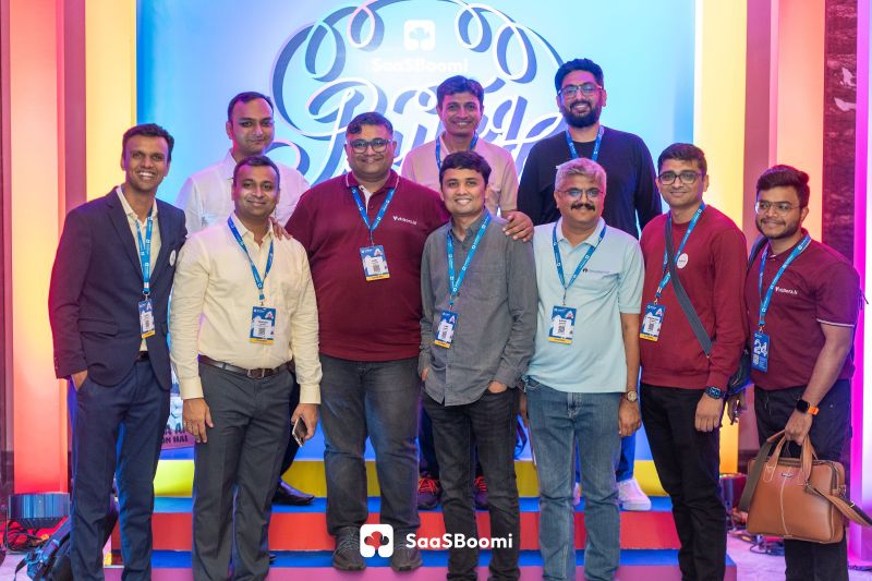 Group of men in lanyards posing on lit stage steps before a colourful SaaSBoomi backdrop at a conference.