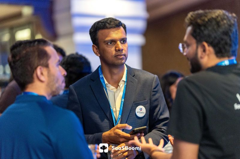 Man in navy blazer holding a phone talks with two other men at a busy conference venue.