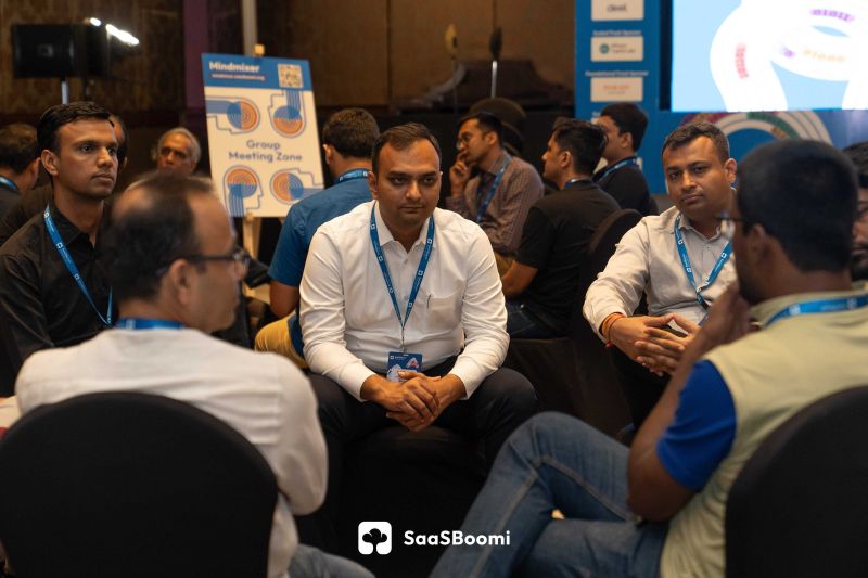 Men seated in a circle listen during a group discussion beside a 'Group Meeting Zone' sign at a conference.