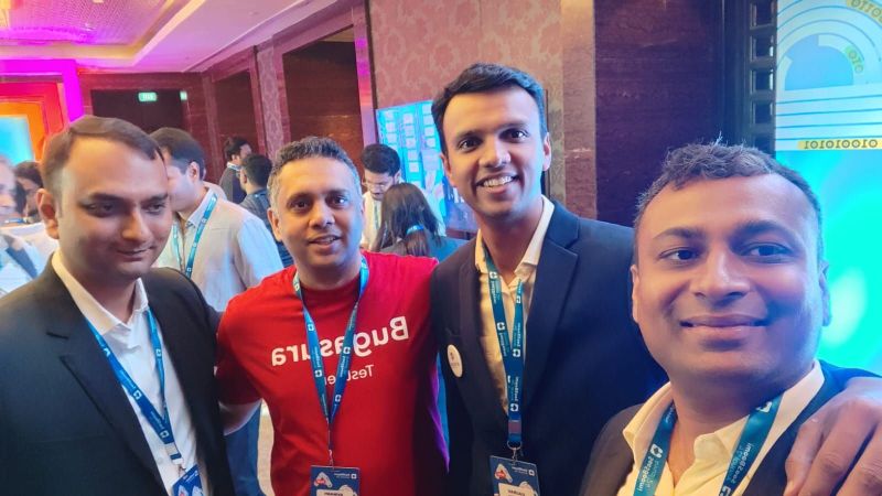 Four men smile together for a close selfie at a conference, one wearing a red Bugasura t-shirt.