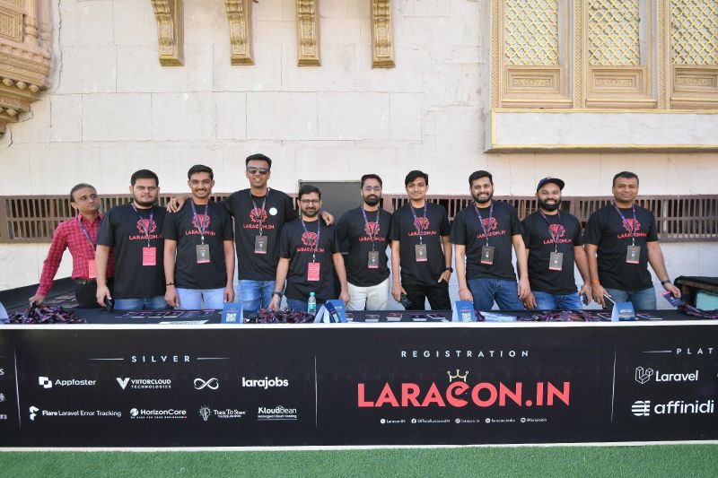 Group of men in black Laracon.IN T-shirts standing behind a registration desk covered with sponsor logos.