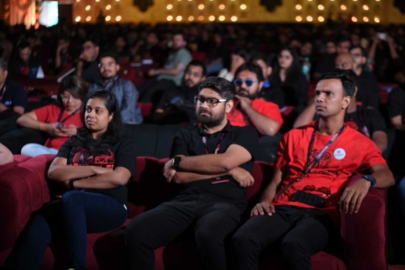 Attendees in red and black T-shirts watch a session from red seats in a dimly lit, packed auditorium.