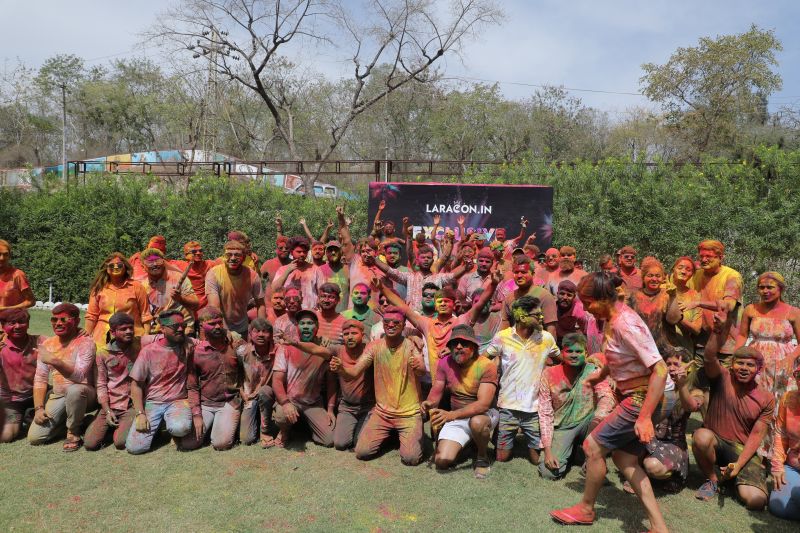 Big crowd covered in bright Holi colours poses and cheers on a lawn in front of a Laracon.IN banner.