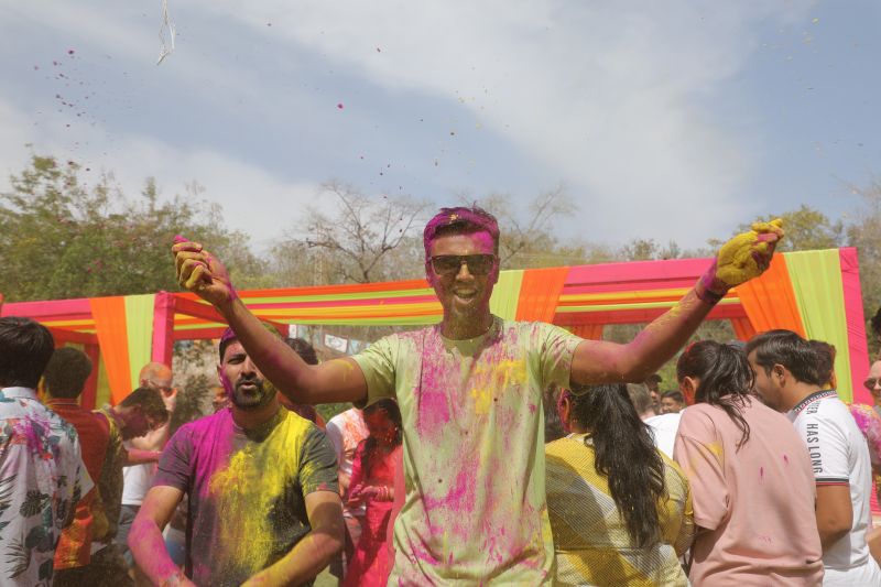 A man smeared with pink and yellow colour raises both arms as colour flies during an outdoor Holi celebration.