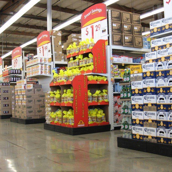 Supermarket aisle endcaps with Smart Savings price signs beside stacked cases of Corona Extra beer.