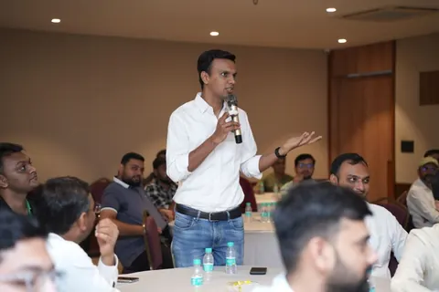 A man in a white shirt speaks into a microphone while standing among seated attendees at round tables.