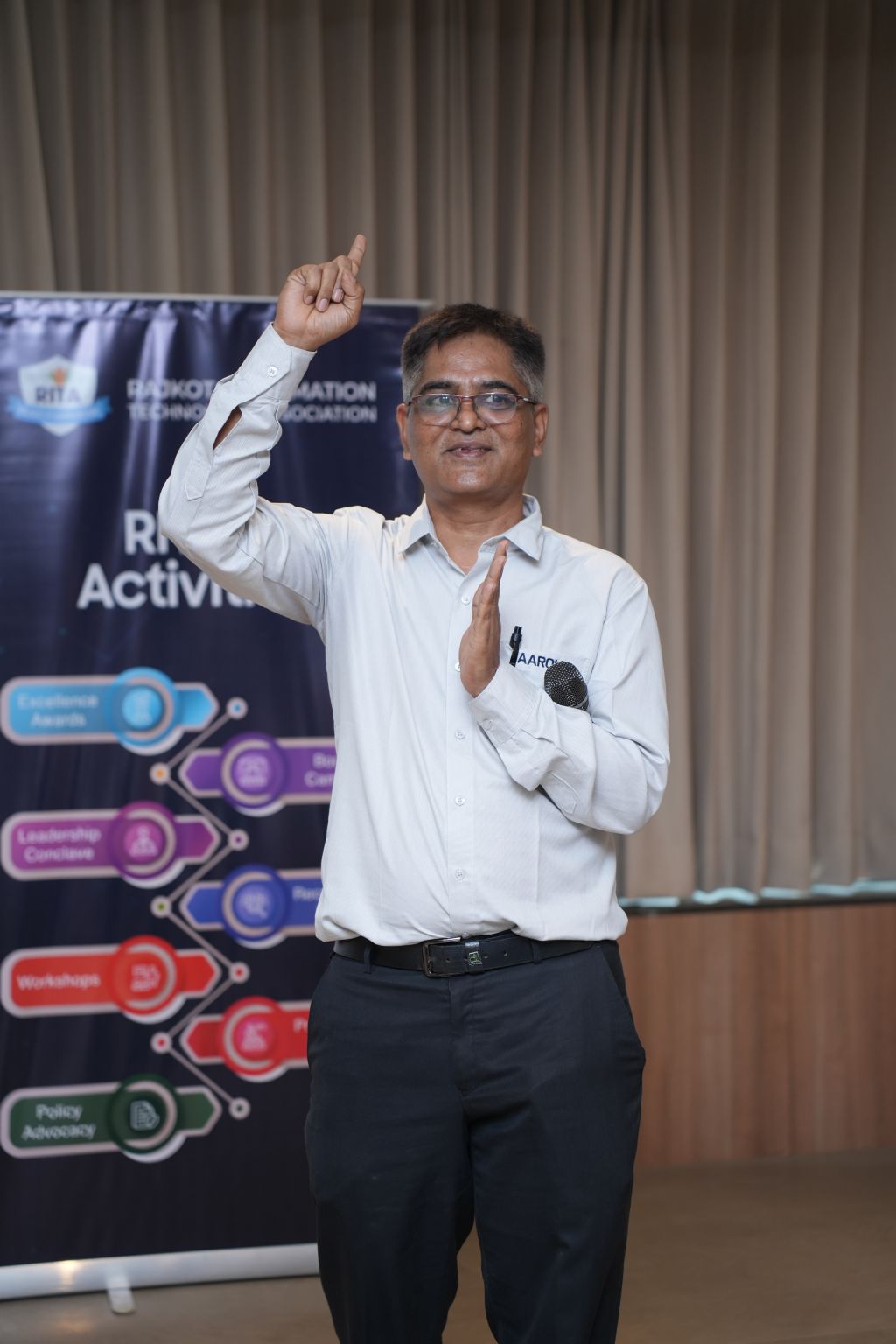 A man with glasses holds a microphone and gestures upward in front of a Rajkot Information Technology Association banner.