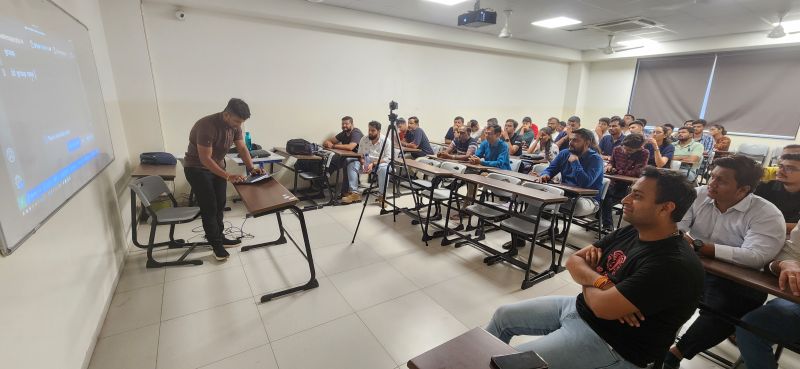 A man stands with a laptop before rows of attendees seated at desks in a classroom during a session.