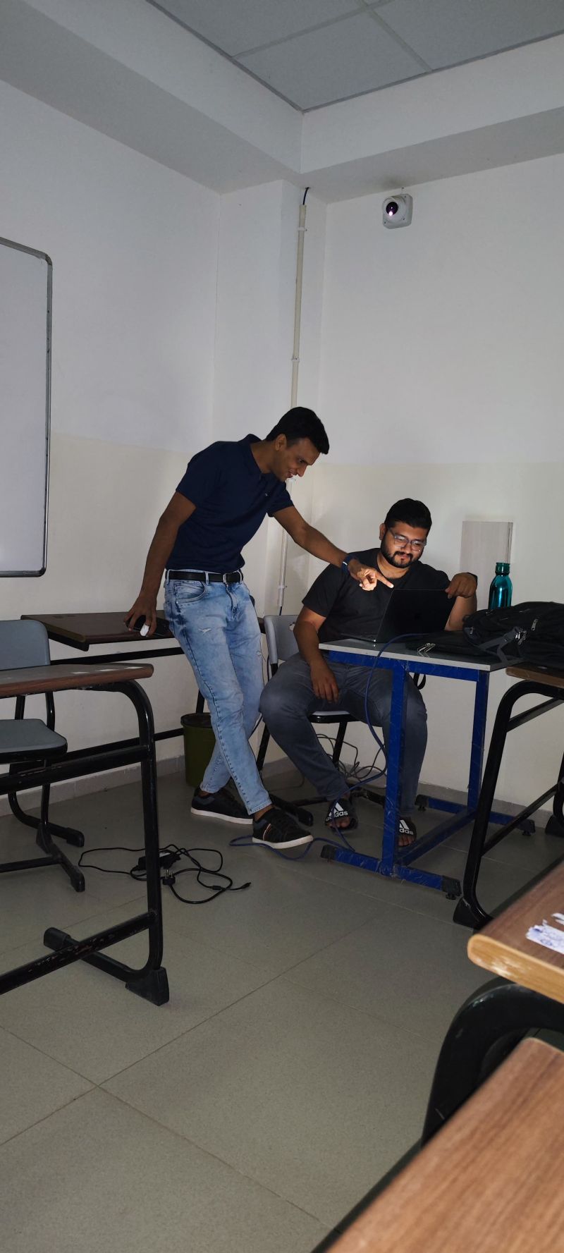 A man points at a laptop screen while another man sits at a desk in a classroom.