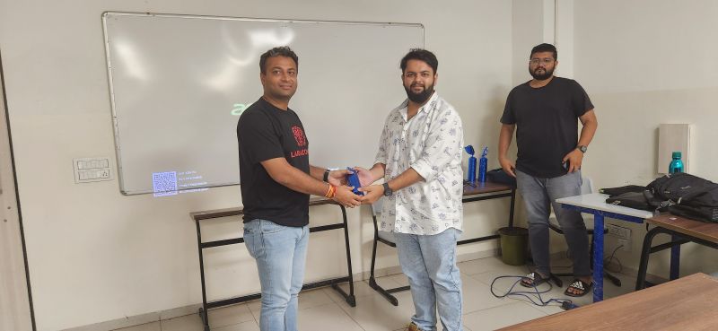 Two men exchange a small blue item while a third man stands nearby in a classroom.
